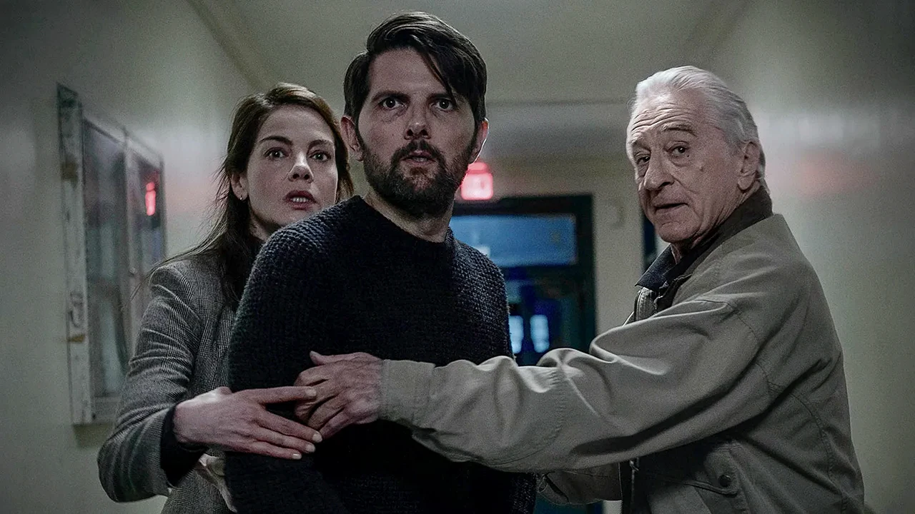 Os atores Robert De Niro, Adam Scott e Michelle Monaghan com expressões assustadas no corredor mal iluminado de uma casa.