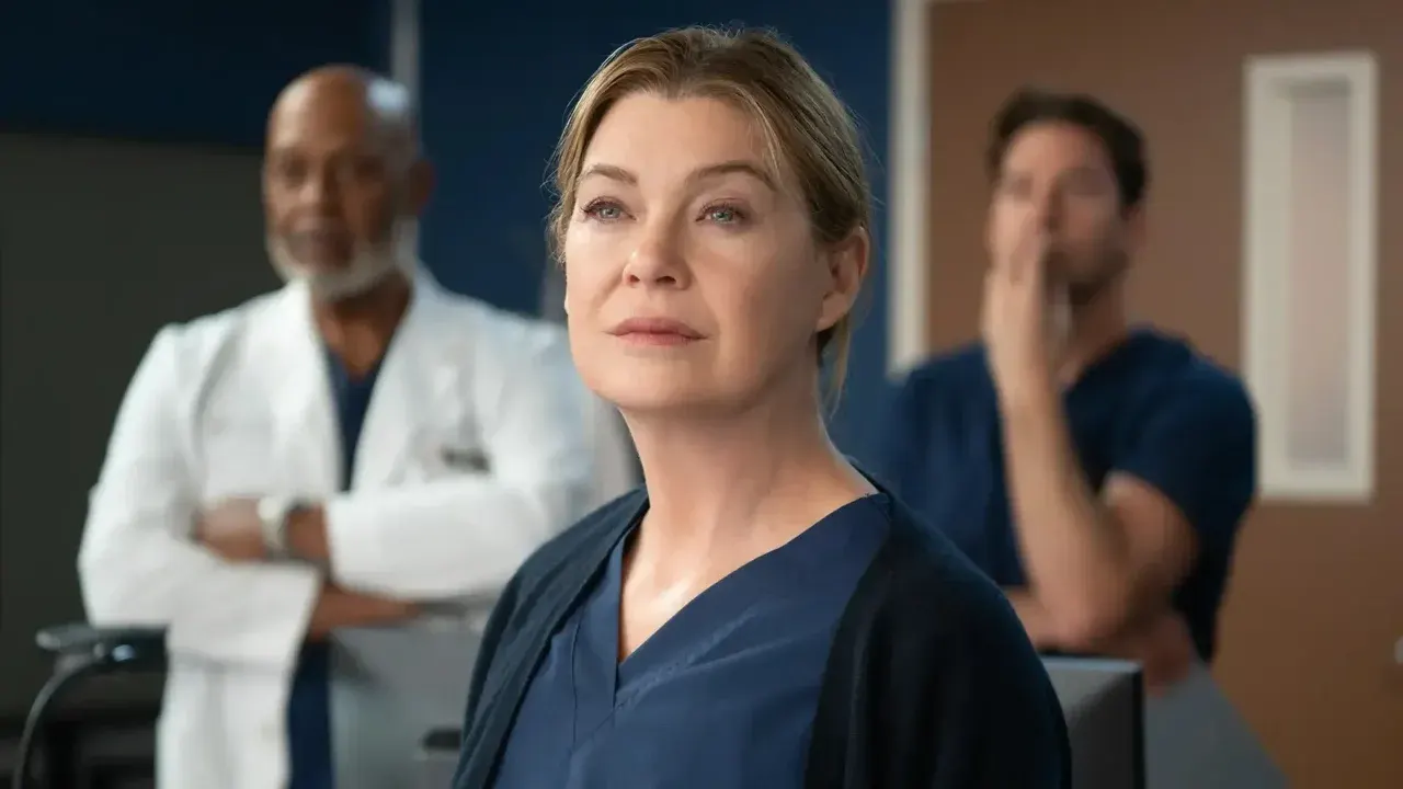 A atriz Ellen Pompeo em primeiro plano com olhar sério, vestindo uniforme médico azul e cardigã preto, com dois outros médicos ao fundo desfocados.