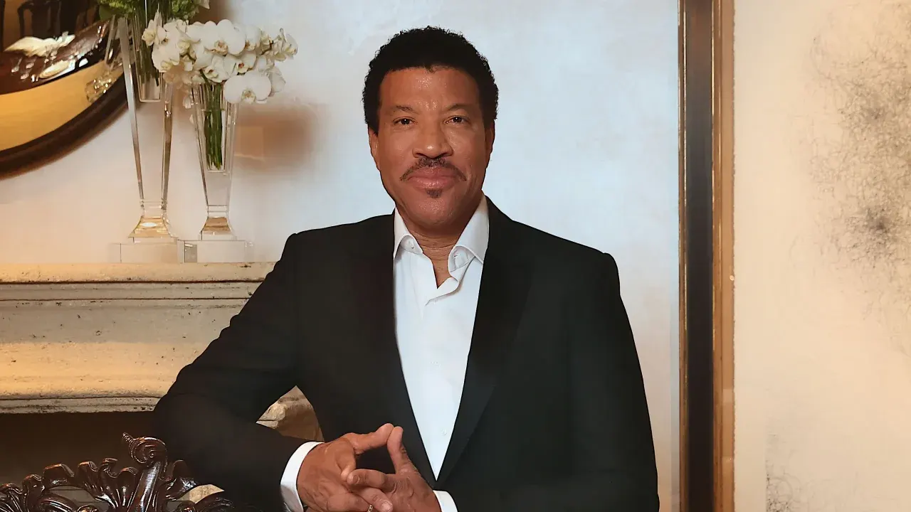 O cantor Lionel Richie vestindo paletó preto e camisa branca, posando com leve sorriso e mãos entrelaçadas em um ambiente elegante.