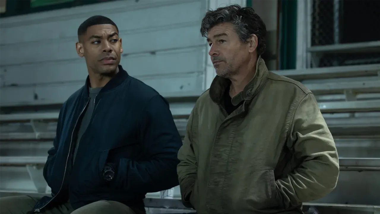Aaron Pierre e Kyle Chandler sentados lado a lado em uma arquibancada com expressões sérias na série Lanternas da DC.