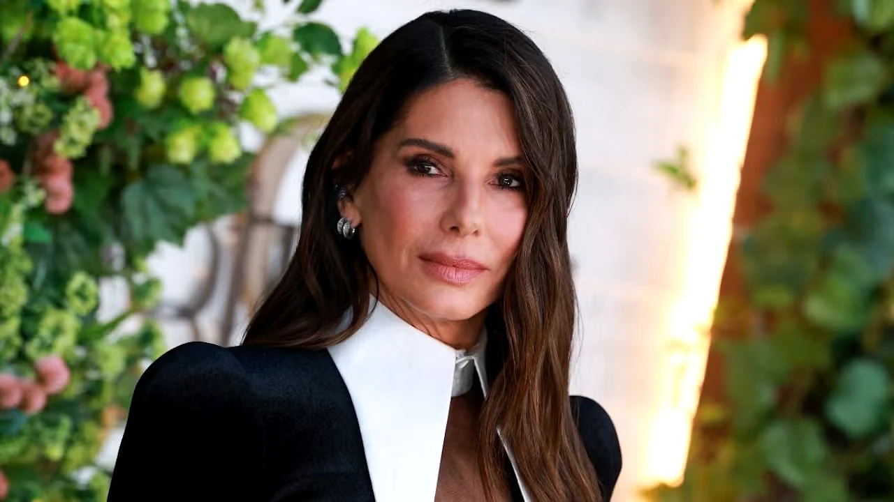 A atriz Sandra Bullock posa levemente sorridente com um vestido preto e gola branca em um evento com fundo de folhagens verdes.