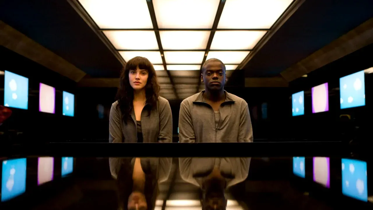 Daniel Kaluuya e Jessica Brown Findlay com expressões sérias emDaniel Kaluuya e Jessica Brown Findlay sentados com roupas cinzas e olhares sérios em sala escura cercada por telas, no episódio de Black Mirror.