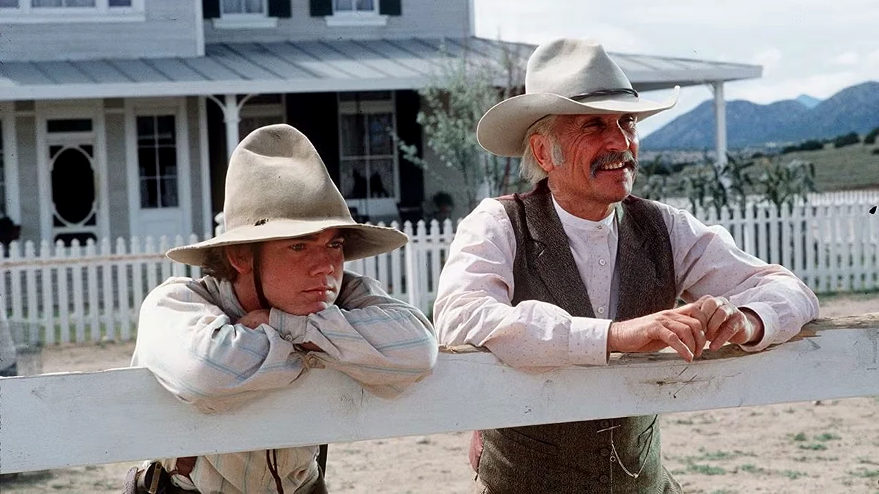 Dois atores caracterizados com roupas de época e chapéus de faroeste encostados em uma cerca branca no set da minissérie Lonesome Dove